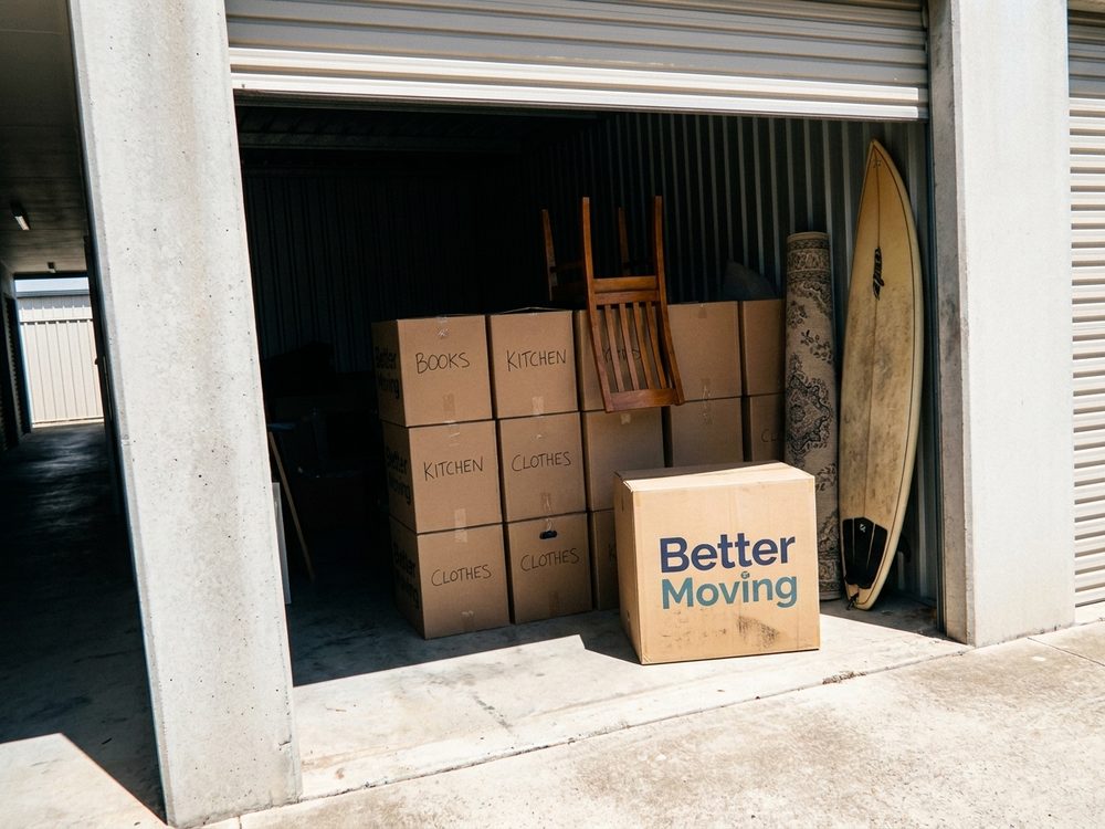 A storage unit with cartons stacked head high, a bike hooked on the wall and a surfboard on its edge.